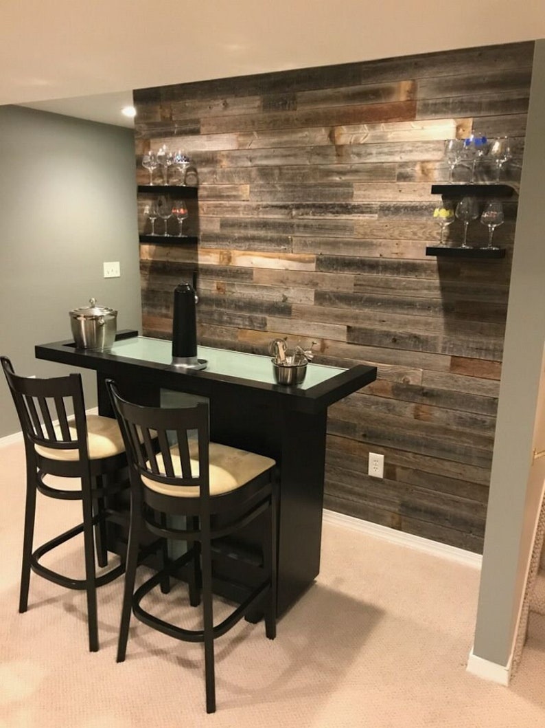 K&ouml;nnte beinhalten: Eine schwarze Bar mit zwei Barhockern und einer Glasplatte. Die Bar steht vor einer Wand mit wiederverwendeten Holzdielen. An der Wand befinden sich zwei Regale mit Weingl&auml;sern.