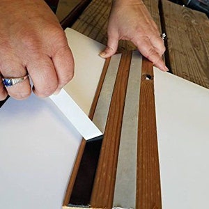 K&ouml;nnte beinhalten: Eine Person klebt transparentes Klebeband auf die R&uuml;ckseite eines Holzbildrahmens. Das Klebeband wird am Rand des Rahmens angebracht, und die Person gl&auml;ttet es mit den Fingern.