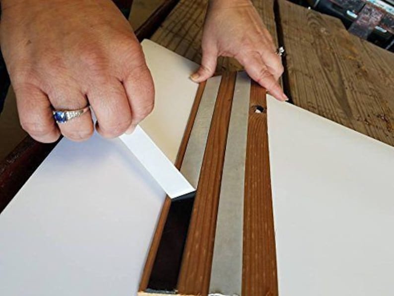 K&ouml;nnte beinhalten: Eine Person klebt transparentes Klebeband auf die R&uuml;ckseite eines Holzbildrahmens. Das Klebeband wird am Rand des Rahmens angebracht, und die Person gl&auml;ttet es mit den Fingern.