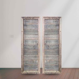 May include: Two rustic wooden doors with a weathered gray finish. Each door is framed with light-colored wood, creating a contrasting border. The doors are made of horizontal planks, giving them a textured appearance. The doors are standing upright against a white wall.