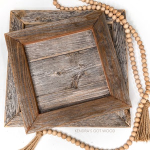 May include: Two wooden picture frames with a rustic, weathered finish, stacked on top of each other. A strand of wooden beads with tassels hangs over the frames. The text "Kendra's Got Wood" is visible on the bottom frame.