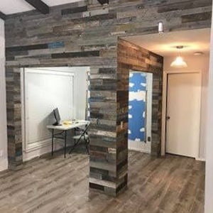 K&ouml;nnte beinhalten: Eine Wand mit Wandverkleidung aus wiederverwendetem Holz in Graut&ouml;nen, Blau und Braun. Die Verkleidung ist vertikal angebracht und erzeugt einen rustikalen und modernen Look. Die Verkleidung wird verwendet, um einen Raumteiler zu schaffen, der zwei Bereiche des Raumes trennt.