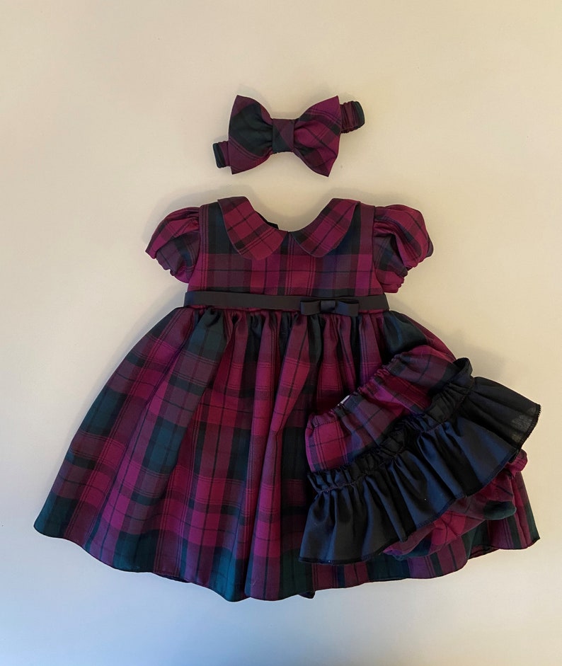 May include: A plaid dress with a black bow and matching headband. The dress has a black belt and a ruffled black underskirt.