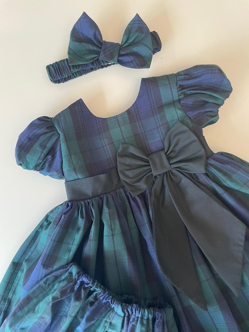 Peut inclure: Une robe &agrave; carreaux verts et bleus avec un n&oelig;ud noir et un bandeau assorti. La robe a des manches bouffantes et une jupe ample.