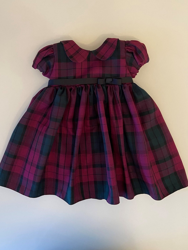 May include: A red and green plaid dress with a black sash and a Peter Pan collar. The dress has short sleeves and a full skirt.