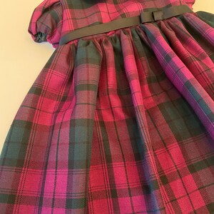 May include: A pink and black plaid dress with a black ribbon and bow at the waist. The dress has a Peter Pan collar and short puffed sleeves.