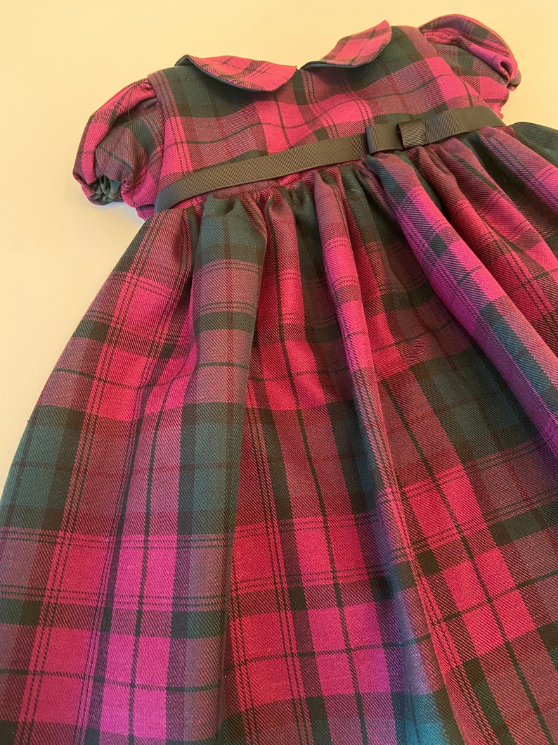 May include: A pink and black plaid dress with a black ribbon and bow at the waist. The dress has a Peter Pan collar and short puffed sleeves.