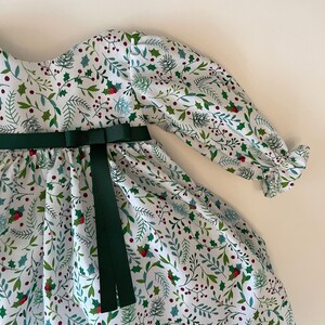 Op de afbeelding: Een witte jurk met een groene bloemenprint en een groen lint geknoopt in de taille. De jurk heeft lange mouwen met ruches aan de manchetten.