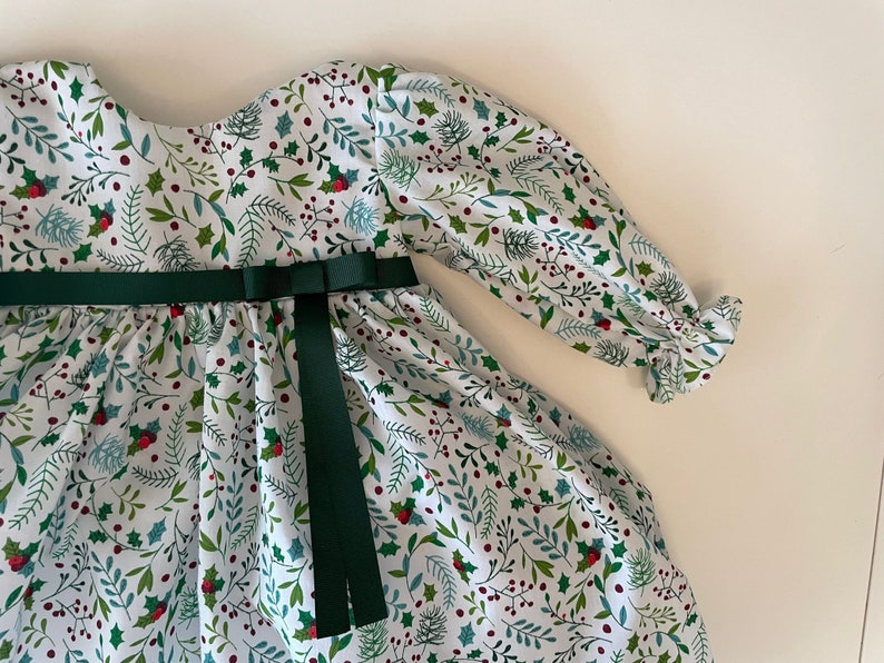 Op de afbeelding: Een witte jurk met een groene bloemenprint en een groen lint geknoopt in de taille. De jurk heeft lange mouwen met ruches aan de manchetten.