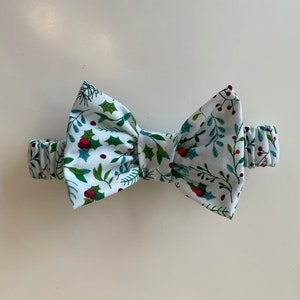 Op de afbeelding: Een witte strik met een groen en rood kerststerrenpatroon. De strik is gemaakt van stof en heeft een elastisch band.