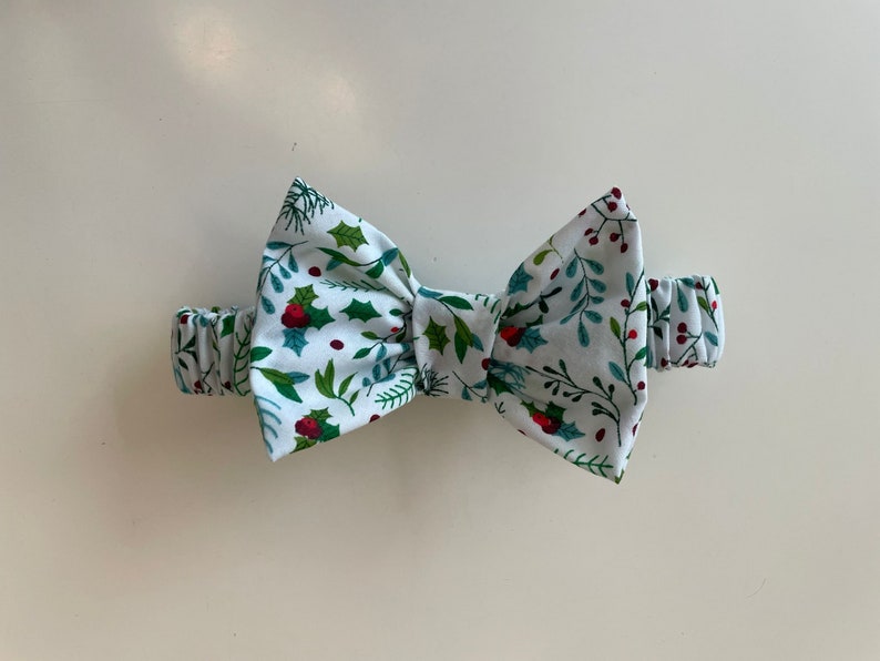 Op de afbeelding: Een witte strik met een groen en rood kerststerrenpatroon. De strik is gemaakt van stof en heeft een elastisch band.