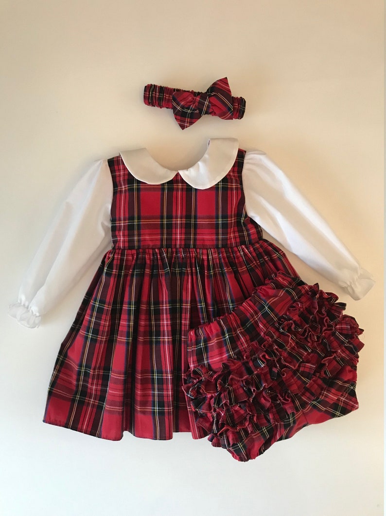 May include: A red and black plaid dress with a white Peter Pan collar and long sleeves. The dress is paired with a matching red and black plaid diaper cover with ruffles. A red and black plaid headband with a bow is included.