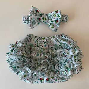 Op de afbeelding: Witte babybloomers met een groene en rode hulstprint en meerdere ruches. Een bijpassend hoofdband met strik is inbegrepen.
