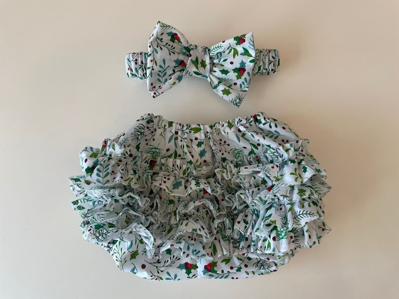 Op de afbeelding: Witte babybloomers met een groene en rode hulstprint en meerdere ruches. Een bijpassend hoofdband met strik is inbegrepen.