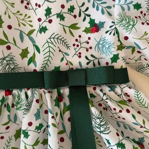 Op de afbeelding: Een witte jurk met een groene en rode bloemenprint. De jurk heeft een groene band die in een strik is geknoopt bij de taille.