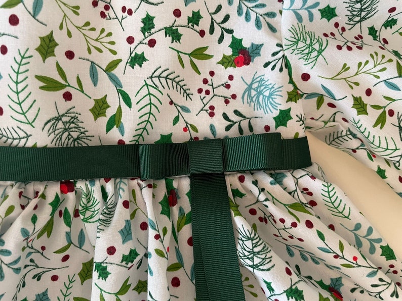 Op de afbeelding: Een witte jurk met een groene en rode bloemenprint. De jurk heeft een groene band die in een strik is geknoopt bij de taille.
