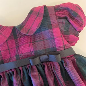 May include: A pink and black plaid dress with a Peter Pan collar and a black ribbon bow at the waist.