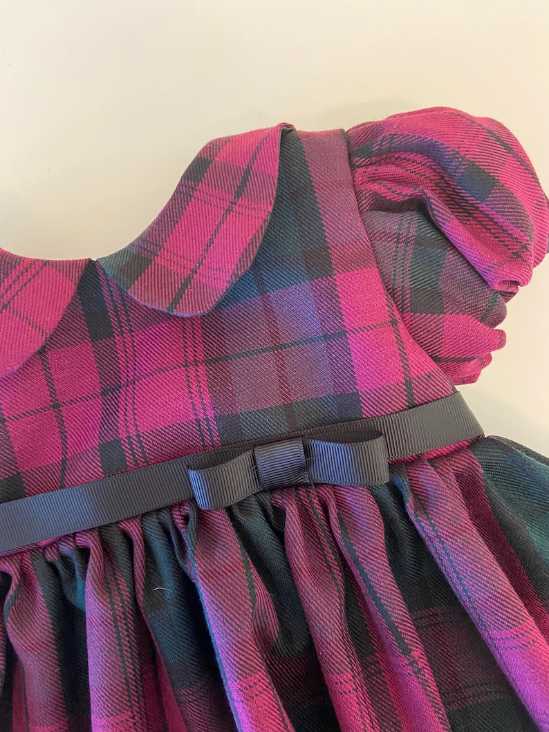 May include: A pink and black plaid dress with a Peter Pan collar and a black ribbon bow at the waist.