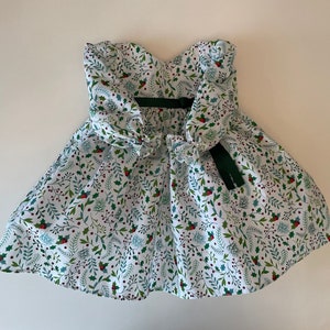 Op de afbeelding: Een witte jurk met een groen en rood bloemenpatroon. De jurk heeft een strapless halslijn en een strik op de rug.