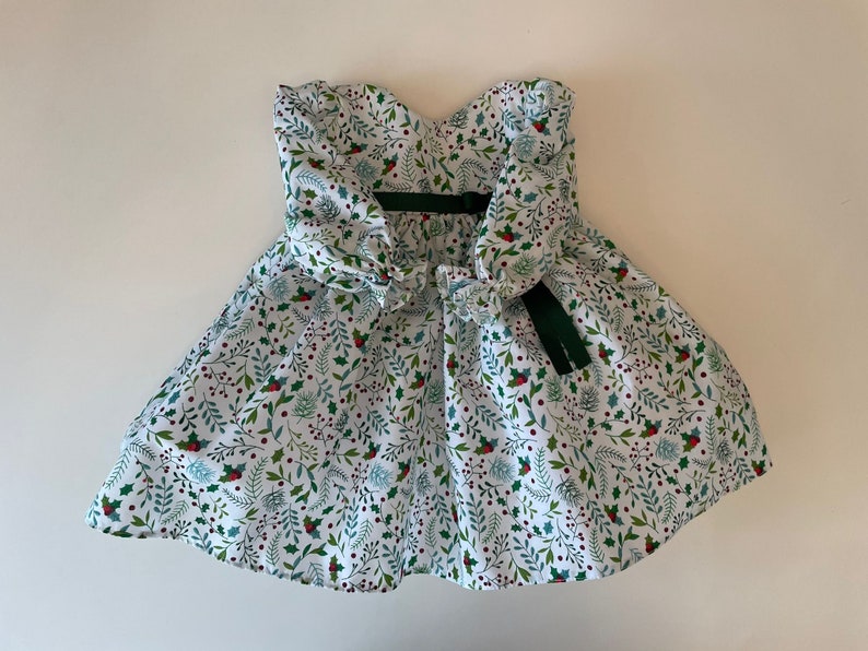 Op de afbeelding: Een witte jurk met een groen en rood bloemenpatroon. De jurk heeft een strapless halslijn en een strik op de rug.