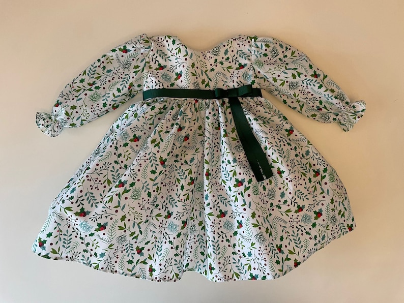 Op de afbeelding: Een witte jurk met een groen en rood hulstpatroon. De jurk heeft lange mouwen met gepofte schouders en een groen lint dat in de taille is geknoopt.