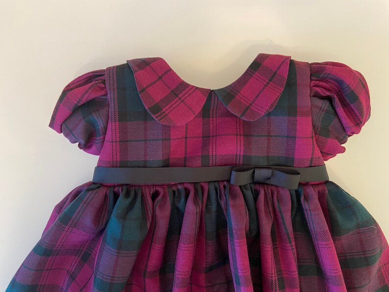 May include: A plaid dress with a black ribbon and bow. The dress has a Peter Pan collar and puffed sleeves.