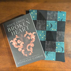 May include: A book titled "ONCE UPON A BROKEN HEART" by Stephanie Garber rests beside a quilted book sleeve. The book cover is dark with a decorative heart design. The book sleeve features a black, teal, and gray plaid pattern with stars and crescent moons.