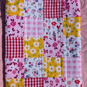 May include: A patchwork fabric item with a variety of floral and gingham patterns in yellow, pink, red, and white. The item is made of squares stitched together, creating a quilt-like appearance. The background is a solid pink.