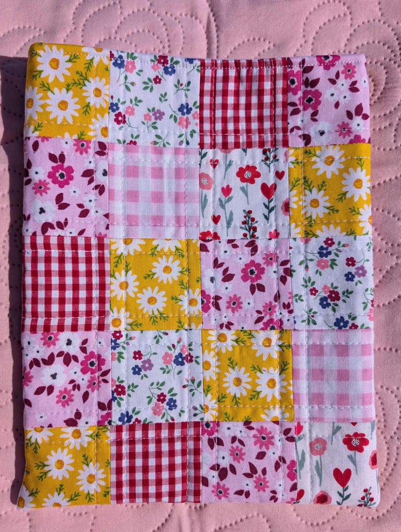 May include: A patchwork fabric item with a variety of floral and gingham patterns in yellow, pink, red, and white. The item is made of squares stitched together, creating a quilt-like appearance. The background is a solid pink.