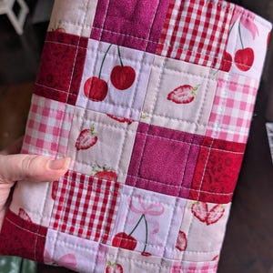 May include: A quilted fabric case featuring a patchwork design with squares of pink, red, and white fabrics. The patterns include cherries, strawberries, and gingham checks. The case is hand-stitched with a pink border.