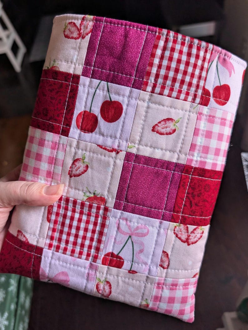 May include: A quilted fabric case featuring a patchwork design with squares of pink, red, and white fabrics. The patterns include cherries, strawberries, and gingham checks. The case is hand-stitched with a pink border.