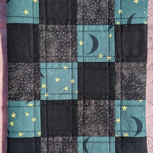 May include: A quilted square featuring a patchwork design with teal, black, and gray squares. Some squares have yellow stars and crescent moons. The item is likely a decorative piece or a small textile item.