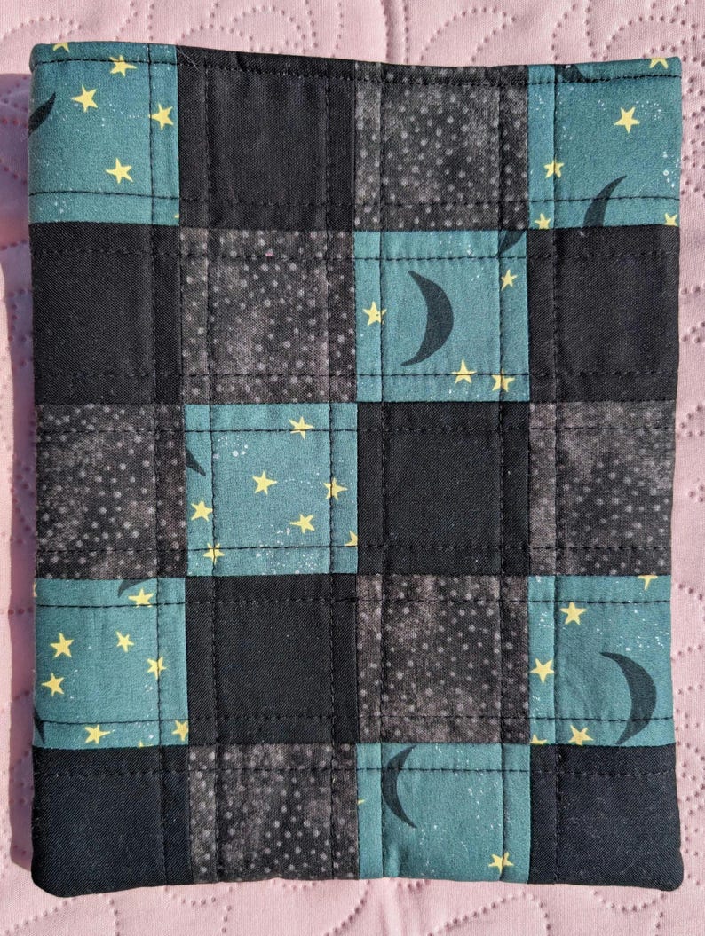 May include: A quilted square featuring a patchwork design with teal, black, and gray squares. Some squares have yellow stars and crescent moons. The item is likely a decorative piece or a small textile item.
