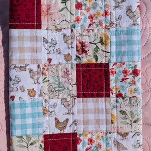 May include: A patchwork fabric item featuring a variety of patterned squares. The squares include floral designs, gingham, and images of chickens. The color palette includes red, white, blue, and beige. The item is likely a small quilt or a similar textile.