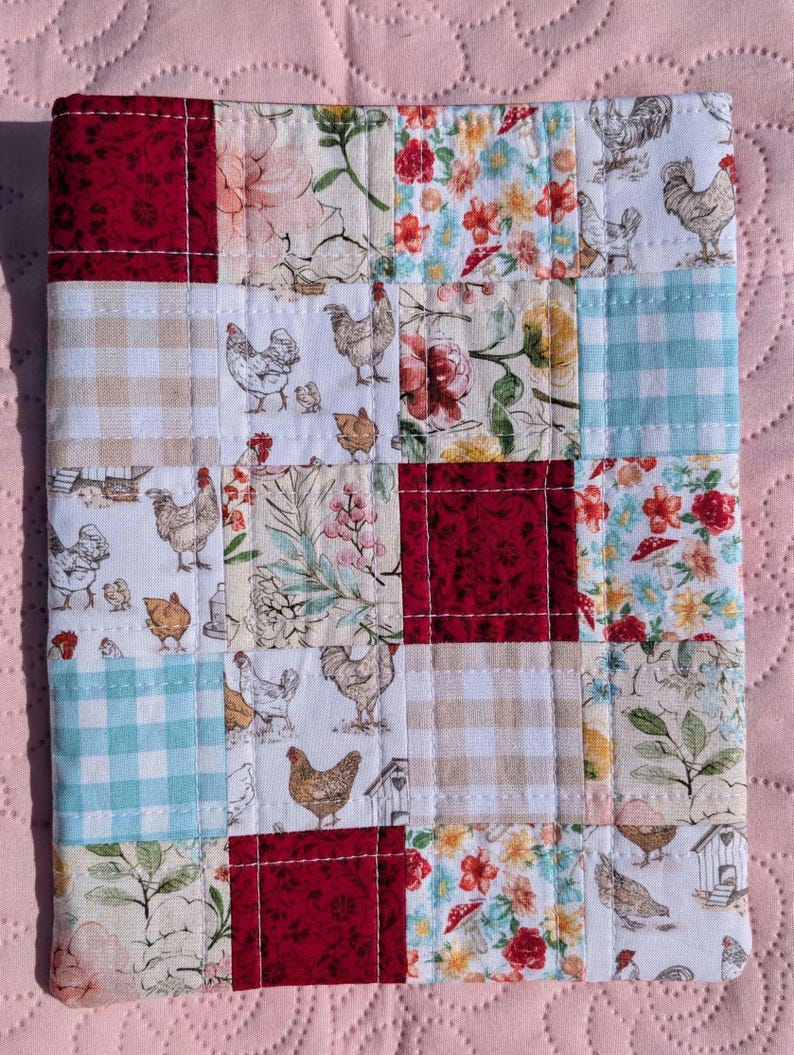 May include: A patchwork fabric item featuring a variety of patterned squares. The squares include floral designs, gingham, and images of chickens. The color palette includes red, white, blue, and beige. The item is likely a small quilt or a similar textile.