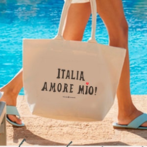 woman walks by the sea in a quaint Italian Harbor holding an off-white  canvas tote bag with the phrase 'ITALIA AMORE MIO' printed in  bold black letters. A small red heart is the dot above the letter 'I'  in the word 'MIO'.