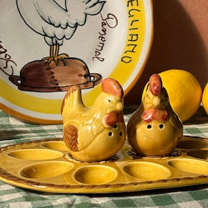 May include: A ceramic plate and egg holder with chicken-themed decor. The plate has a yellow rim and a rooster illustration. Two chicken-shaped salt and pepper shakers sit on the egg holder. Lemons and text are also visible.