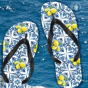 Op de afbeelding: Een paar blauwe en witte slippers met een citroenmotief. De slippers hebben zwarte bandjes en zijn geplaatst tegen een blauwe waterachtergrond.