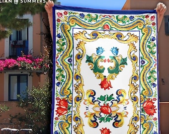 May include: A white beach towel with a colorful Italian-inspired design. The design features a blue, yellow, and green floral pattern with red accents. The towel has a blue border.