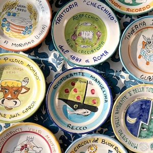 Op de afbeelding: Een verzameling kleurrijke, handgeschilderde keramische borden met diverse Italiaanse restaurant- en voedselgerelateerde ontwerpen. Elk bord heeft unieke illustraties, tekst en levendige kleuren, die een rustieke, ambachtelijke stijl laten zien.
