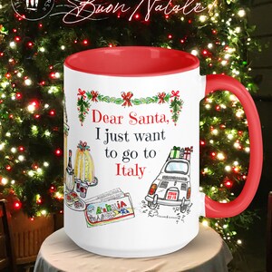 Op de afbeelding: Een witte keramische mok met een rode rand en handvat. De mok heeft een kleurrijke illustratie van een Fiat-auto met kerstcadeaus op het dak, een instapkaart voor een vlucht naar Italië en de tekst "Lieve Kerstman, ik wil gewoon naar Italië". De mok is 15 ounce.
