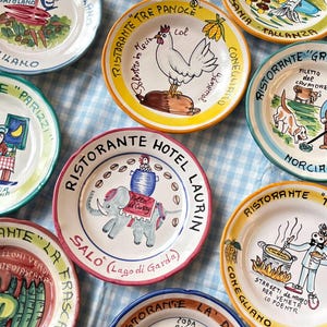 May include: A collection of round ceramic plates featuring hand-painted illustrations of animals, food, and restaurant names. Each plate has a unique colorful rim and detailed scenes including an elephant, a chicken, and various culinary motifs.