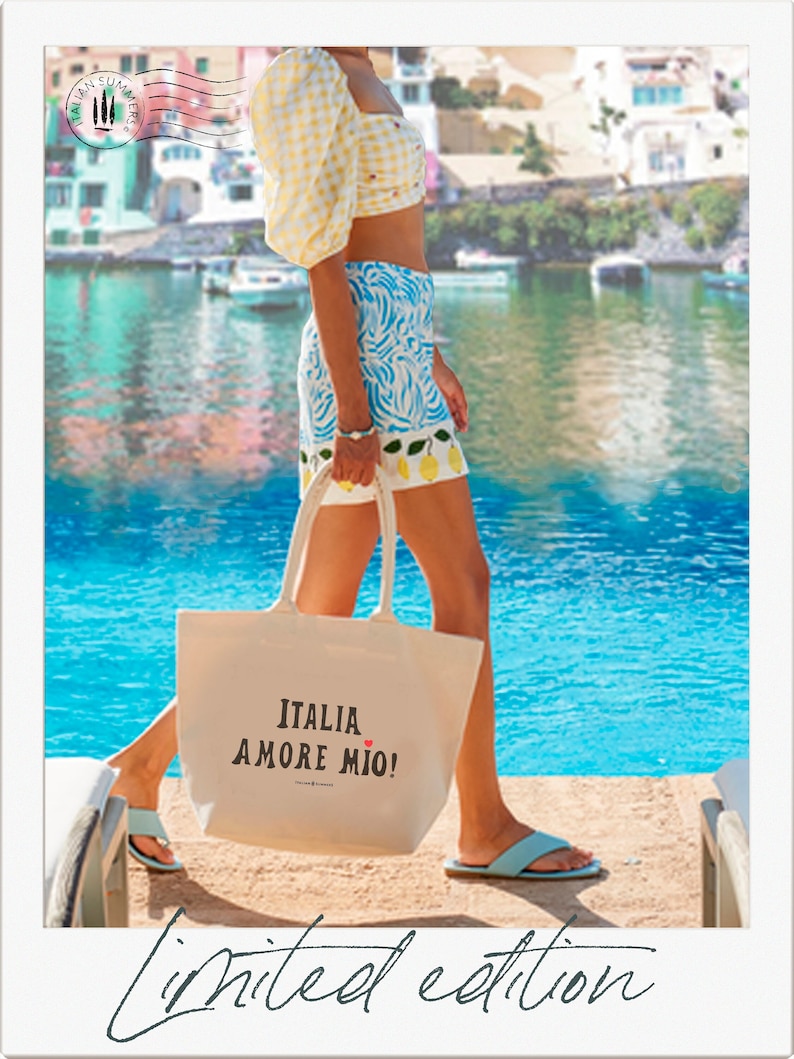 woman walks by the sea in a quaint Italian Harbor holding an off-white  canvas tote bag with the phrase 'ITALIA AMORE MIO' printed in  bold black letters. A small red heart is the dot above the letter 'I'  in the word 'MIO'.