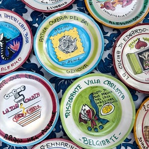 Op de afbeelding: Een verzameling handbeschilderde keramische borden met kleurrijke ontwerpen. Elk bord heeft unieke illustraties, waaronder vissen, eten en tekst in het Italiaans. De borden zijn gerangschikt op een blauw-wit patroon tafelkleed.