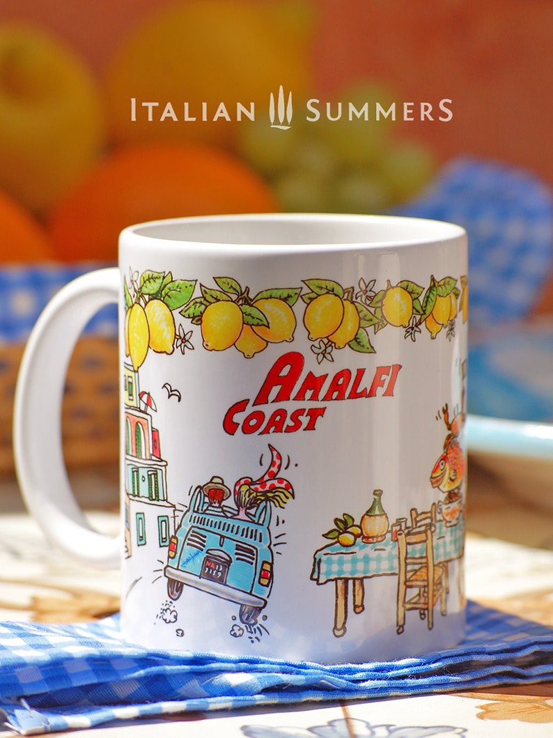 May include: White ceramic mug with a colorful illustration of a car driving through a town with the text "Amalfi Coast" and lemons on a vine. The mug is on a blue and white checkered tablecloth.