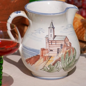 May include: White ceramic jug with a blue rim and a painted scene of a church with a red roof and green plants. The jug has a handle and is on a white tablecloth.