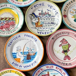 May include: A collection of hand-painted ceramic plates with colorful illustrations and text. Designs include a cow, a fish, a fisherman, and various Italian restaurant scenes. The plates have a rustic, artisanal aesthetic, with each plate featuring unique artwork and text.