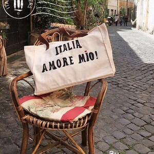 An off-white  canvas tote bag with the phrase 'ITALIA AMORE MIO' printed in  bold black letters. A small red heart is the dot above the letter 'I'  in the word 'MIO'.