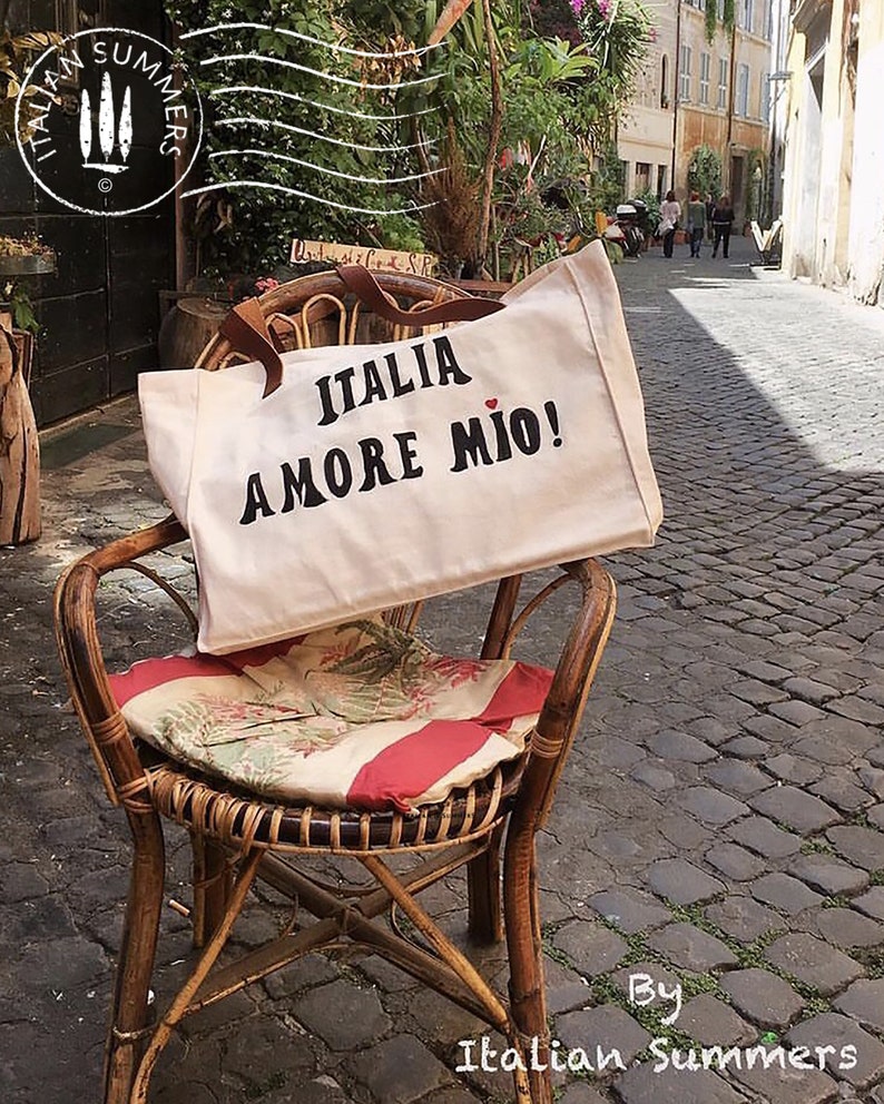 An off-white  canvas tote bag with the phrase 'ITALIA AMORE MIO' printed in  bold black letters. A small red heart is the dot above the letter 'I'  in the word 'MIO'.