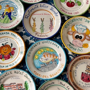 Può includere: Una collezione di nove piatti rotondi in ceramica, ognuno caratterizzato da illustrazioni dipinte a mano di cibo, animali o edifici, accompagnati da vari nomi di ristoranti italiani e località scritti lungo i bordi.